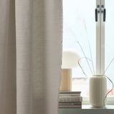 Shared living room scene with grey VÄXELBRUK curtain, small lamp on books, tall white vase with branches.