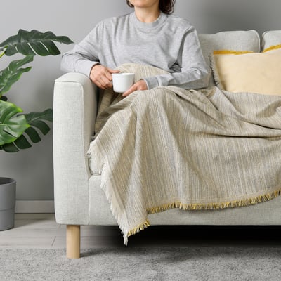 Person sitting on light grey IKEA sofa with a large, light-colored throw blanket with fringe. They hold a white mug. A large plant is to the side.