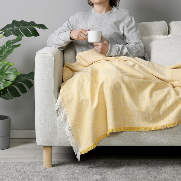 Person on sofa with VÄXELBRUK blanket drinking coffee, plant, rug.