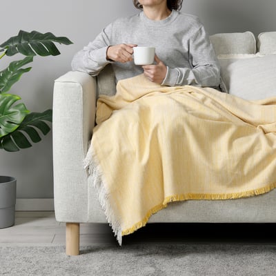 Person on sofa with VÄXELBRUK blanket drinking coffee, plant, rug.