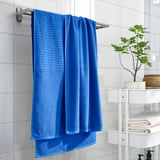 Bright blue VÅGSJÖN towel hangs on silver rack above white shelving with folded towels and green plant, contrasting white tiled wall.