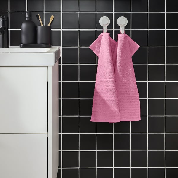 Bright pink VÅGSJÖN towel hangs on wall, contrasting dark tiles, two white hooks. Soft cotton, medium size, folded, modern bathroom.
