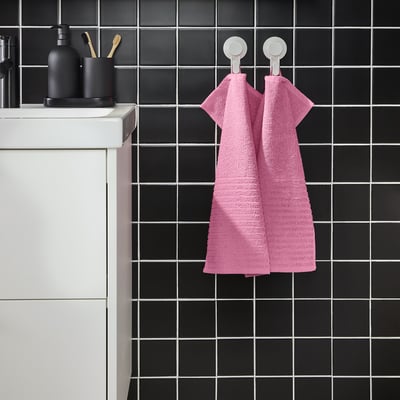 Bright pink VÅGSJÖN towel hangs on wall, contrasting dark tiles, two white hooks. Soft cotton, medium size, folded, modern bathroom.
