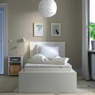 Minimalist white bedroom: small white bed against gray wall with two framed pictures, a round pendant light, and a small white nightstand with a lamp.