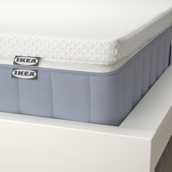 Image an IKEA mattress with a removable, textured top layer, sitting on a white base. Labels show branding on the side.