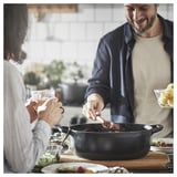 In a man and woman enjoy cooking together using a VARDAGEN black enamelled cast iron pan on a wooden table with food and drinks nearby.