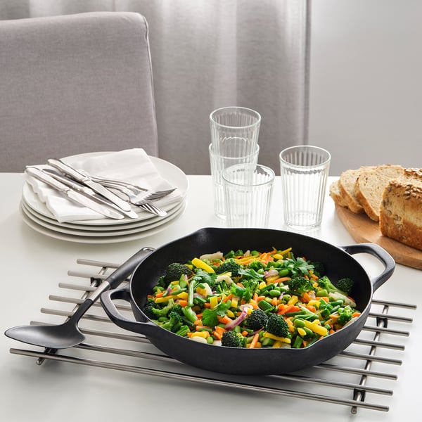 Cast iron skillet with stir-fried vegetables on a trivet, plates, napkins, and cups nearby.