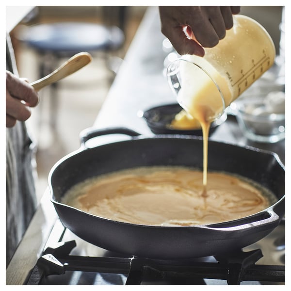 A person is pouring yellow liquid from a measuring cup into a black frying pan on a stove.