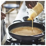 A person is pouring yellow liquid from a measuring cup into a black frying pan on a stove.