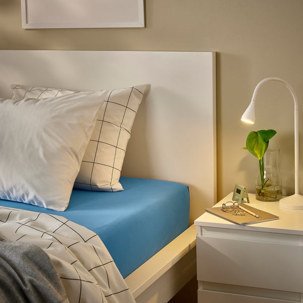 A neatly made bed with VÅRVIAL blue cotton jersey bedding. A white lamp, keys, and a plant sit on a small nightstand beside it.
