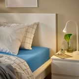 A neatly made bed with VÅRVIAL blue cotton jersey bedding. A white lamp, keys, and a plant sit on a small nightstand beside it.