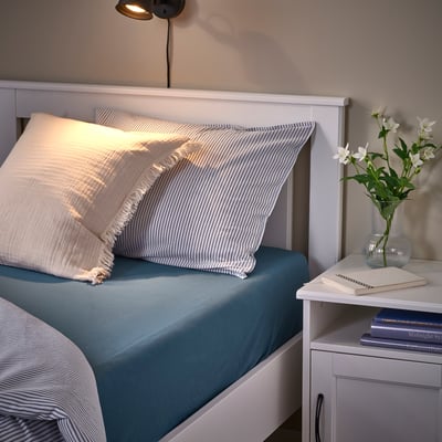 Cozy bed with VÅRVIAL headboard, blue spread, striped pillows. Nightstand with vase, books, lamp.