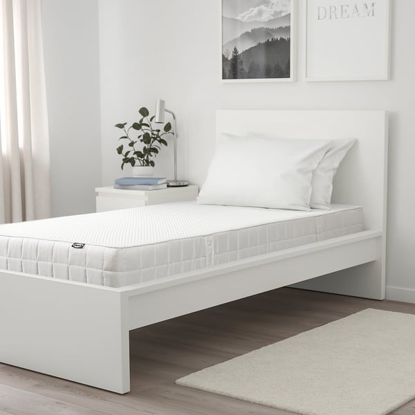 Serene bedroom: minimalist bed with white mattress, two pillows, nightstand holding books, lamp, and plant. Two wall art pieces and rug.