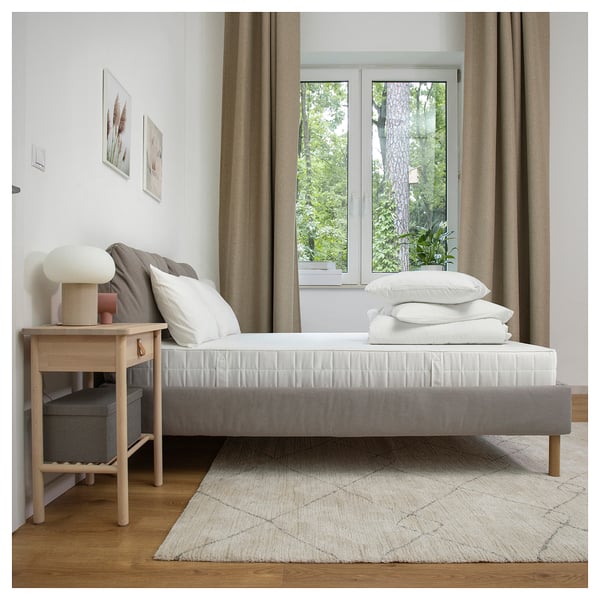 Bedroom with minimalist bed, white pillows, wooden nightstand, lamp, window, curtains, rug, and wall art; cozy and modern.