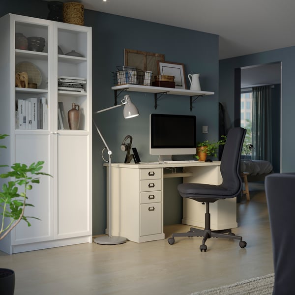 A modern home office setup with a white desk, grey ergonomic chair, and bookshelf, arranged against a green wall.