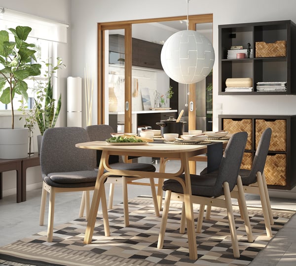 Modern dining area with VEDBO chairs, light wood table, gray cushions, checkered rug, hanging lamp, plants, and shelves with books.