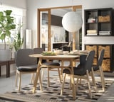 Modern dining area with VEDBO chairs, light wood table, gray cushions, checkered rug, hanging lamp, plants, and shelves with books.