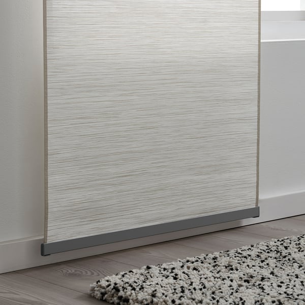 A curtain panel holder attached to a light-colored wooden floor, securing a white-silver patterned curtain, with a textured rug in front.