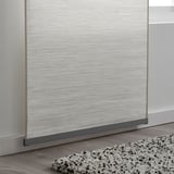 A curtain panel holder attached to a light-colored wooden floor, securing a white-silver patterned curtain, with a textured rug in front.