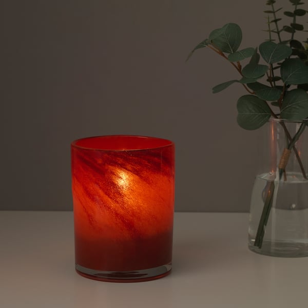 Lit red glass candle holder, emitting warm light. Beside it, a glass vase with green plant.