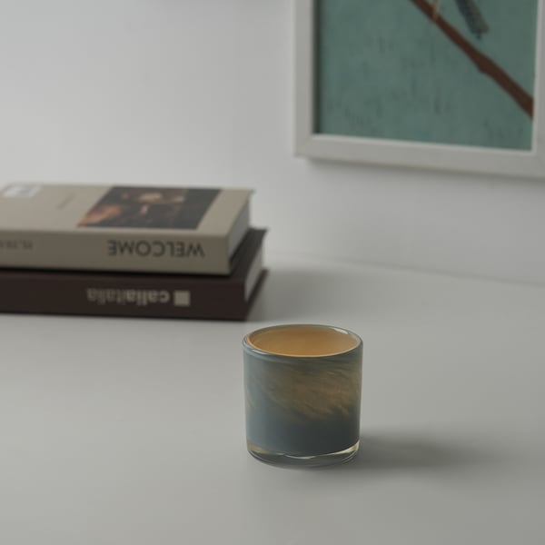 Colorful glass candle holder with green-gold swirls on white surface next to books and abstract painting.