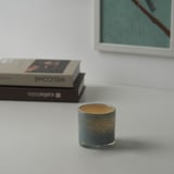 Colorful glass candle holder with green-gold swirls on white surface next to books and abstract painting.