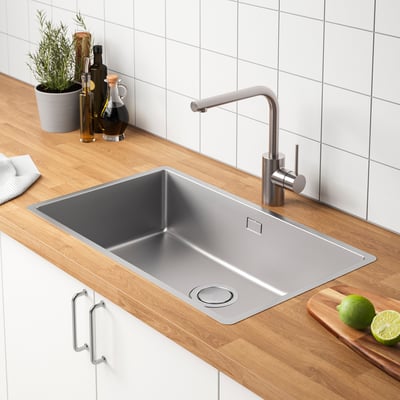 Stainless steel kitchen sink with faucet, set on wooden countertop. Next to a cutting board with limes and adjacent to condiments.