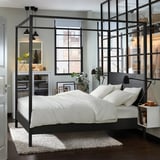 Modern black four-poster bed, centered rug, white bedding, large windows.