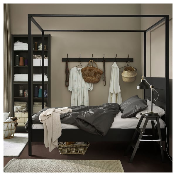 Bedroom with black metal bed, grey bedding, clothes, and baskets. Lamp and basket on bed, shelves with clothes.