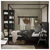 Bedroom with black metal bed, grey bedding, clothes, and baskets. Lamp and basket on bed, shelves with clothes.