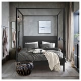 Grey four-poster bed, black headboard, cotton bedding, basket, coats, art, window with curtains.