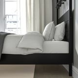 Bed by window, white sheets, black frame. Sunlight streams through curtains.