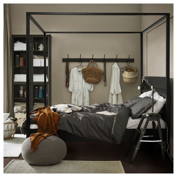 Modern four-poster bed with sleek black frame, high headboard, dark grey linen bedding, black side table, and beige wall hangings.