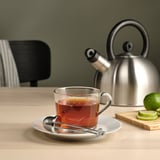 En te-infusionsenhed i et glaskrus med en metalkedel bagved.