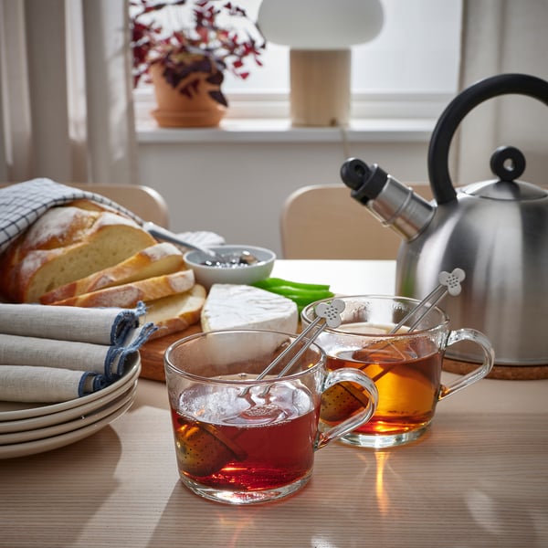 To krus med te-infusionsenheder og te ved siden af brød, marmelade og en tekande på et bord, hvilket tyder på en hyggelig te-tid-opsætning.