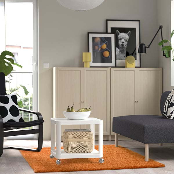 Moderne stue med orange tæppe, hvidt bord, grå sofa, sort-hvid stol, træskab og indrammede billeder.