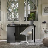 Lille, minimalistisk skrivebord ved navn ALEX, sort med en hvid stol, placeret under store vinduer, omgivet af planter og indretning i et lyst rum.