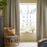 Roligt værelse med lysegrå ANNAKAJSA gardiner, træstol, pude, lampe, plante og bøger ved et stort vindue.