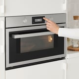 Person indstiller moderne mikrobølgeovn i rent køkken. Finish i rustfrit stål, digitalt display 150, med skarp funktion og afrimningsindstillinger.