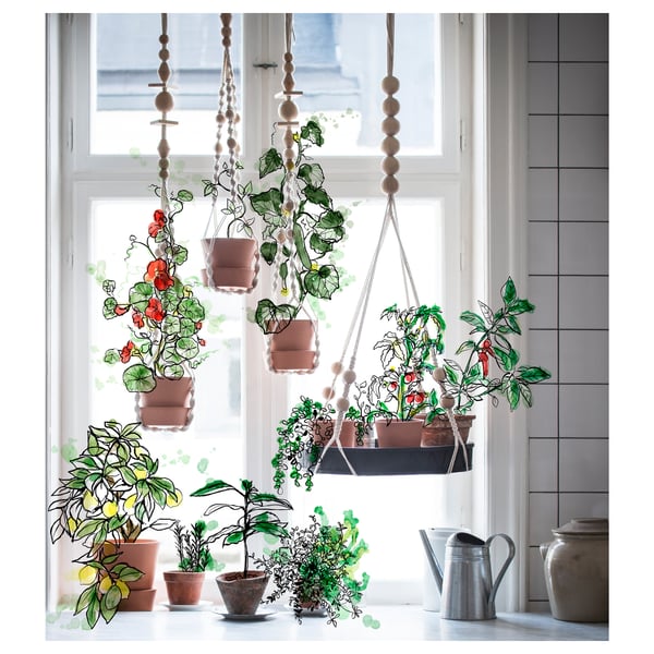 Hængende potteplanter ved vindue med varieret grønt, blomster, jordkrukker, træperler, hvide gardiner og grå vandkande.