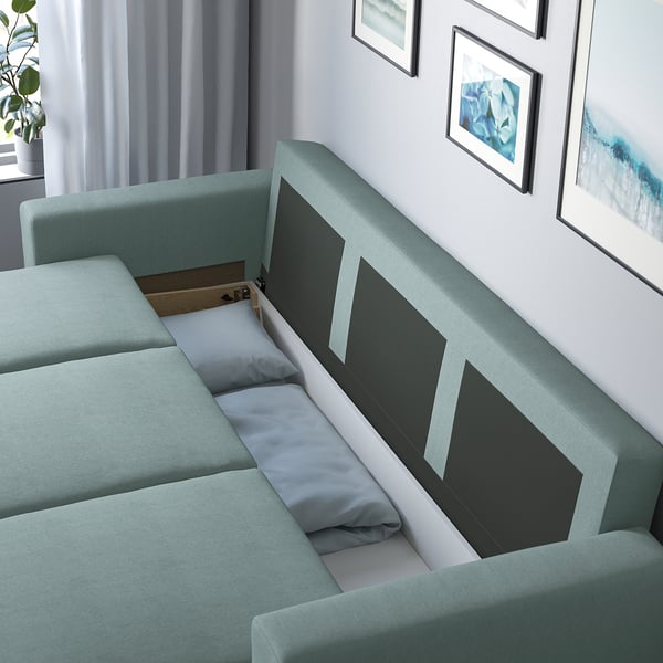 Grøn sovesofa med åben opbevaring, der viser hynder og kasse indeni. Moderne stue med kunst og planter.