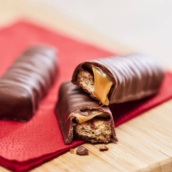Tre chokoladebarer på rød serviet, en skåret op med sprød havrekiks og sej karamel indeni.