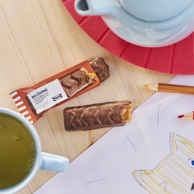 To ikea BELÖNING chokoladebarer på et træbord, den ene pakket ud og bidt, ved siden af kopper med drikkevarer, en rød dækkeserviet og farveblyanter.