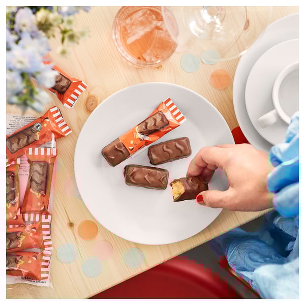 En hånd i en blå handske rækker ud efter en chokoladebar på en hvid tallerken, omgivet af flere barer og indpakninger på et dekoreret træbord.