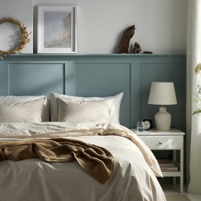 Roligt blåt soveværelse med hvid seng, beige tæppe, puder, sidebord, lampe, bøger og kunst.