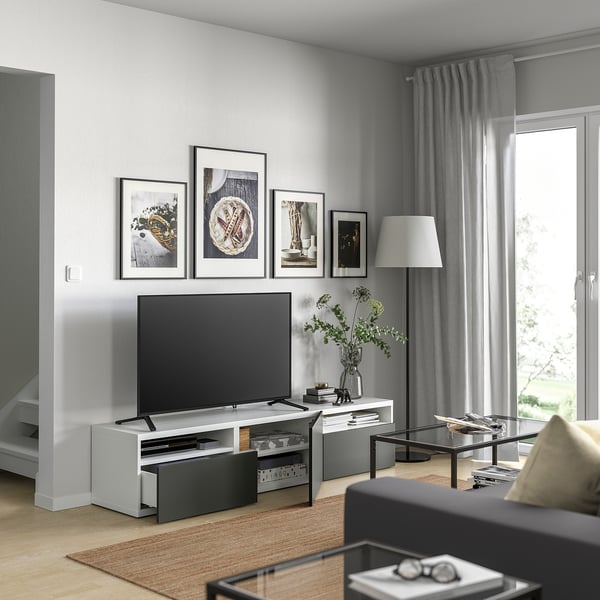 Stue med en BESTÅ hvid tv-bænk, fladskærm, moderne kunst, grå gardiner, stort vindue, tæppe, sofa og sofabord i glas.