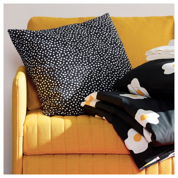 En gul sofa, en sort og hvid prikket pude og en sort klud med hvide blomstermotiver.