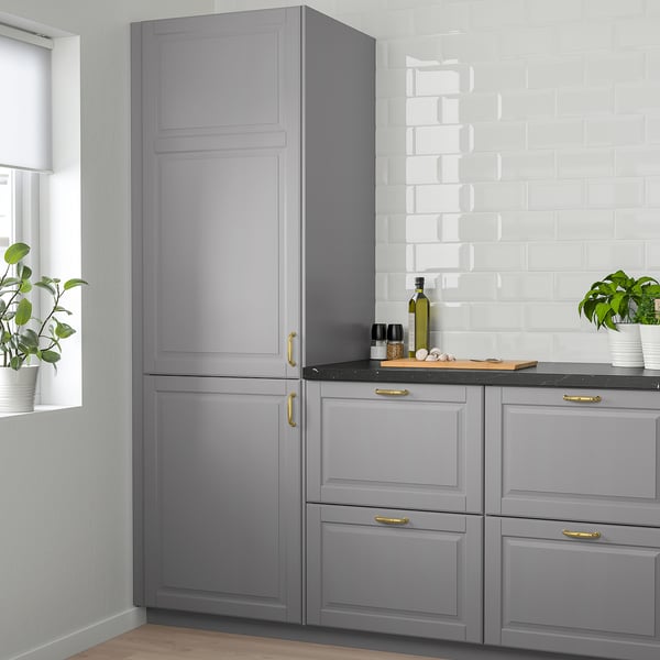 Et højt gråt køkkenskab, højere end nærliggende potteplanter, med et klassisk design med skrå paneler og guldgreb.