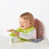 Baby sidder i højstolen, holder et grønt bæger med hvid tud, iført stribet tøj og grøn hagesmæk.