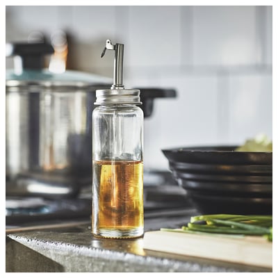 Oliedispenser i klart glas med metaldyse på køkkenbordet i nærheden af skærebræt og grønne løg. Enkelt, moderne design.